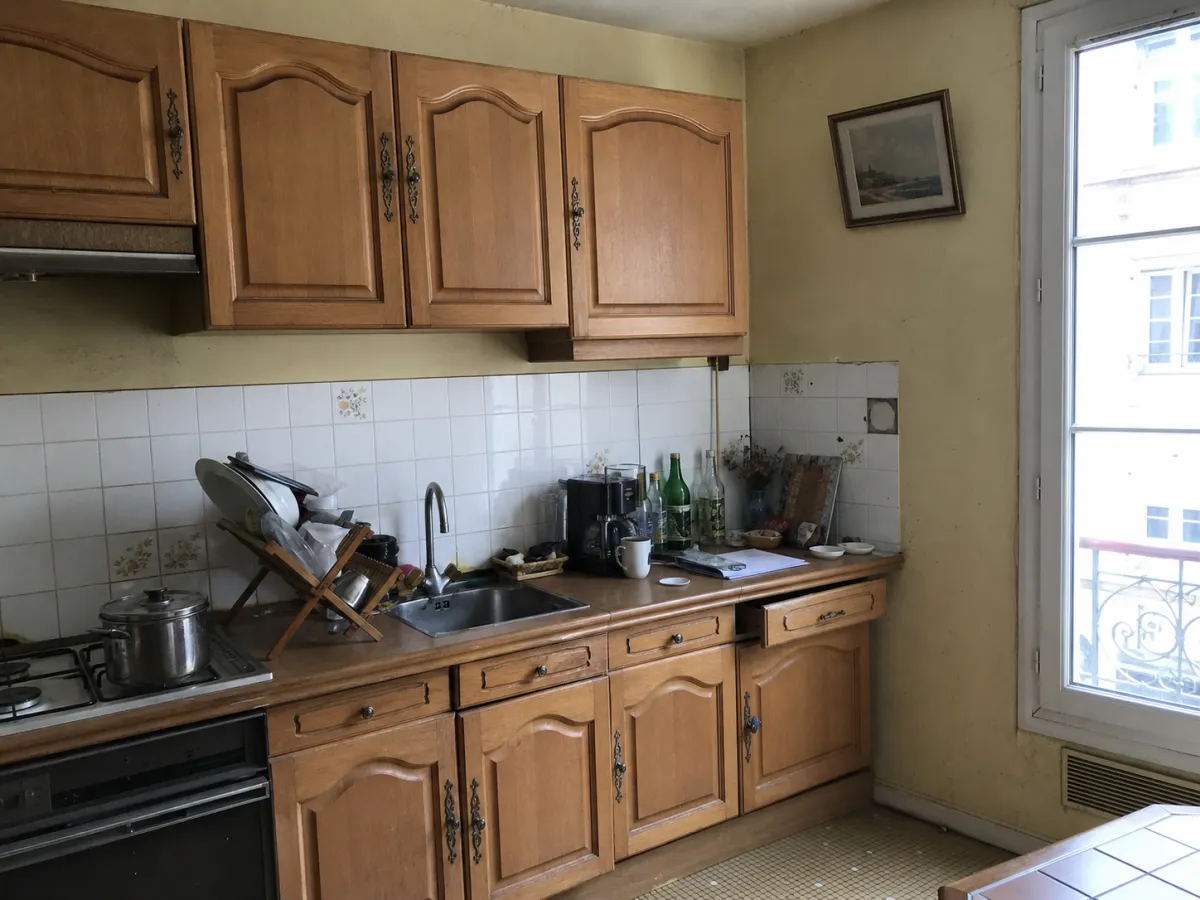 Cuisine avant relooking virtuel : meubles en bois et plan de travail encombré, à côté d’une porte-fenêtre.